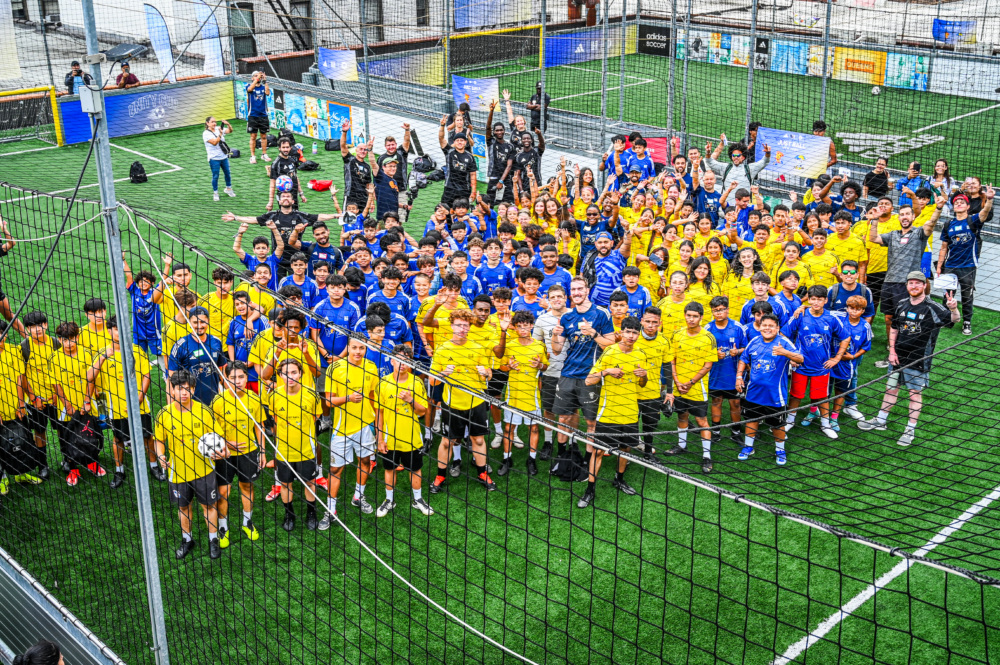 hundreds of kids in blue and yellow jerseys on a dark green turf outdoor soccer field smile up at the camera as they prepare to have fun celebrating the end of the soccer league season. this group is made up of just ball league participants.